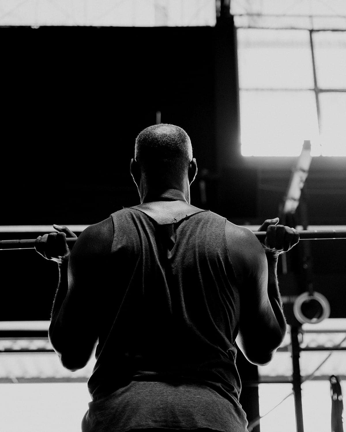 An athlete holds a loaded barbell at the shoulders, facing a tall factory window.