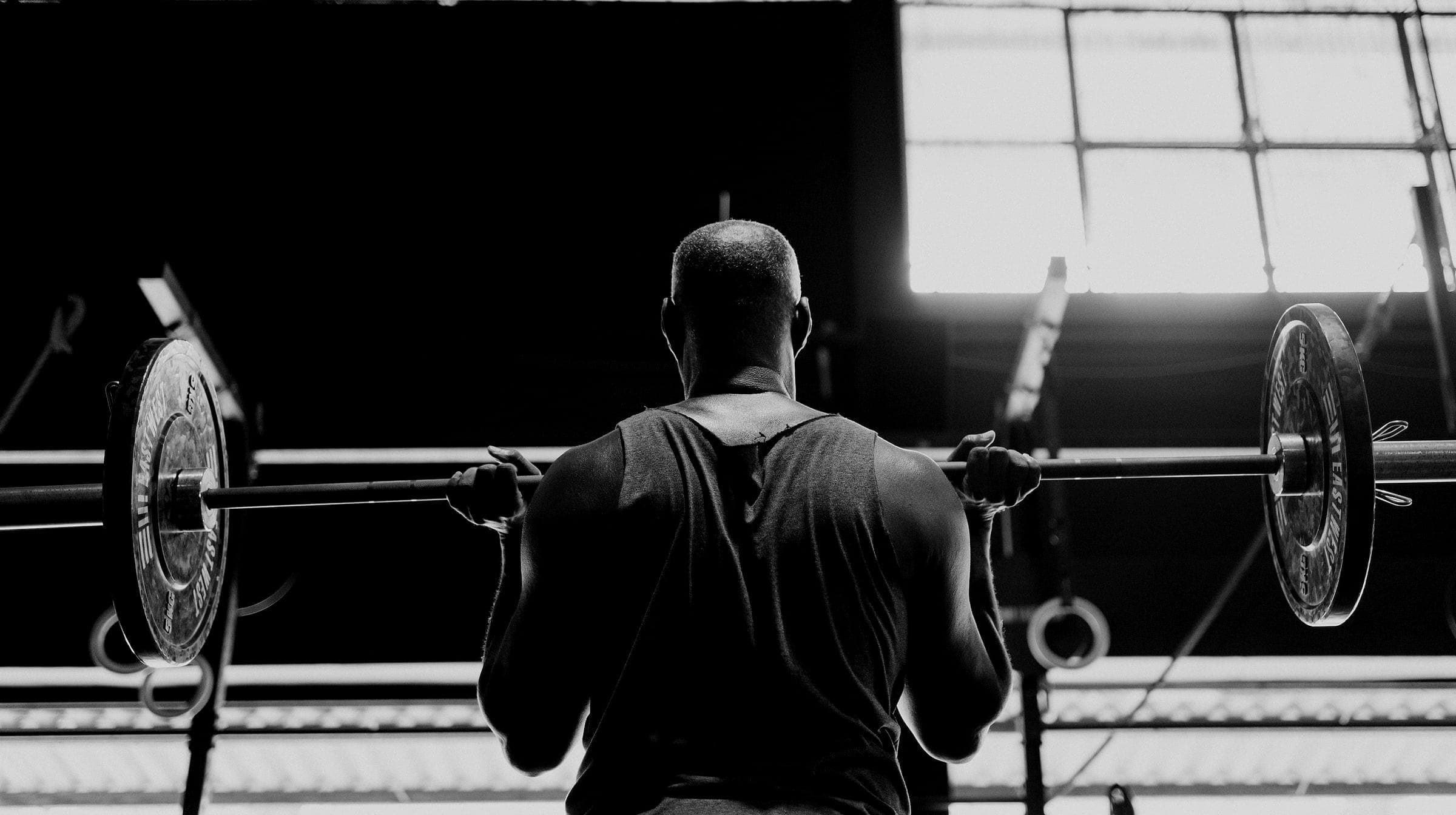 An athlete holds a loaded barbell at the shoulders, facing a tall factory window.