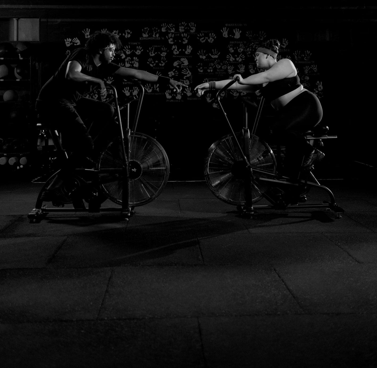 Two riders on air bikes in a dark training room.